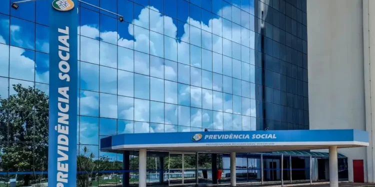 Fachada do prédio do INSS com reflexo do céu e nuvens no vidro, com o letreiro 'Previdência Social' em destaque, indicando o local de atendimento do Instituto Nacional do Seguro Social.