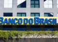 Placa amarela com letras azuis do Banco do Brasil em fachada de prédio moderno cinza com janelas espelhadas