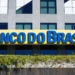 Placa amarela com letras azuis do Banco do Brasil em fachada de prédio moderno cinza com janelas espelhadas