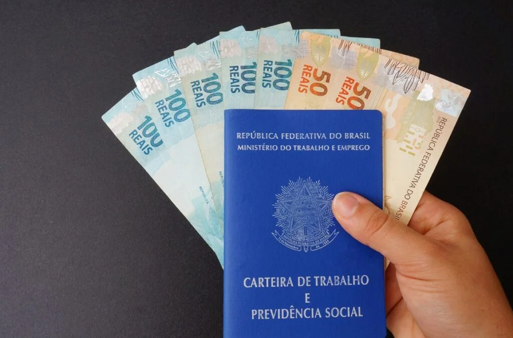 Como solicitar o Seguro-Desemprego em 2026 Pessoa segurando a carteira de trabalho e cédulas de dinheiro, representando o seguro-desemprego.