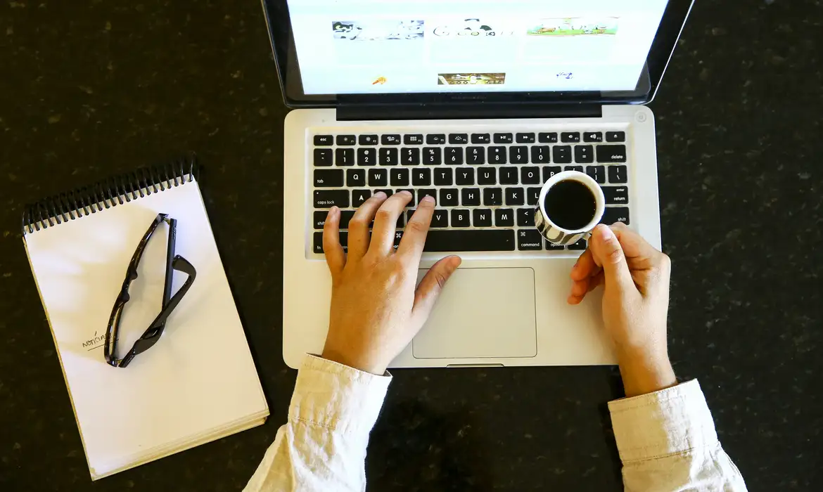 Trabalho remoto com laptop e café na mão Pessoa usando laptop com uma mão no teclado e outra segurando uma xícara de café ao lado de caderno e óculos