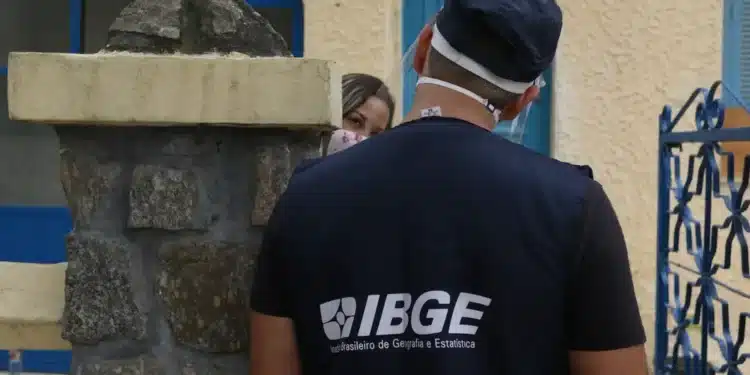Servidor do IBGE com colete azul e logotipo, conversando com uma moradora, ambos usando máscara.