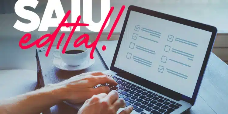 Mãos digitando em notebook com checklist na tela e xícara de café ao lado com texto Saiu Edital