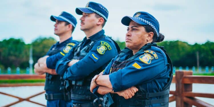 Três guardas municipais de uniforme azul em pé, braços cruzados, ao ar livre