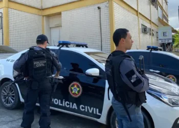 Dois policiais civis armados ao lado de uma viatura com a identificação da Polícia Civil