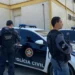 Dois policiais civis armados ao lado de uma viatura com a identificação da Polícia Civil