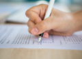 Mão segurando caneta branca e escrevendo em papel com texto impresso sobre mesa de madeira clara