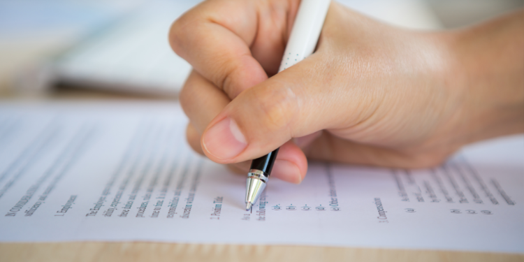 Mão segurando caneta branca e escrevendo em papel com texto impresso sobre mesa de madeira clara