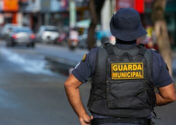 Agente da Guarda Municipal de costas, patrulhando uma rua movimentada em 2026