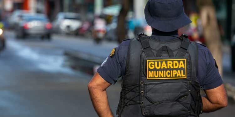 Agente da Guarda Municipal de costas, patrulhando uma rua movimentada em 2026