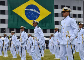 Oficiais da Marinha do Brasil em traje branco durante cerimônia, com grande bandeira do Brasil ao fundo.