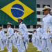 Oficiais da Marinha do Brasil em traje branco durante cerimônia, com grande bandeira do Brasil ao fundo.
