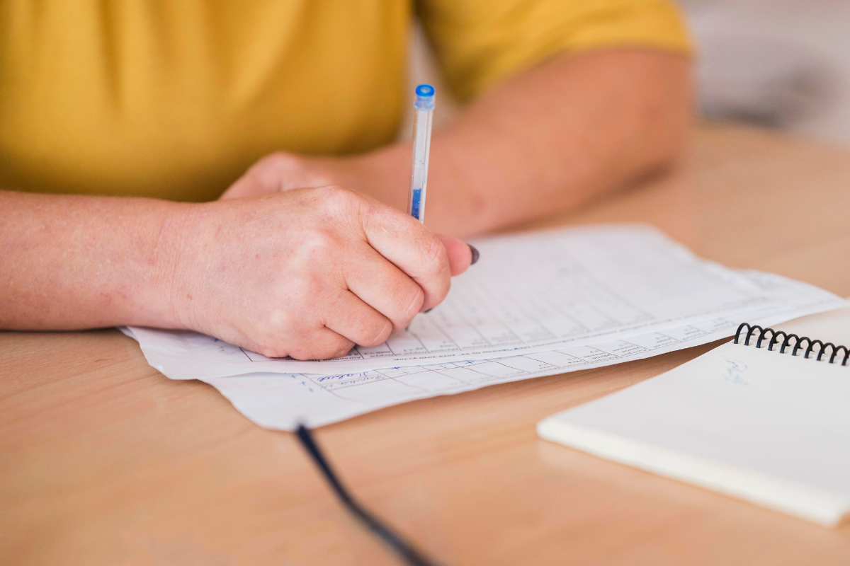 Concursos 2026 Pessoa de camisa amarela preenchendo formulário com caneta azul sobre mesa