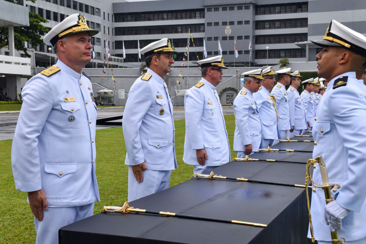 Concursos Marinha Oficiais da Marinha em fila, usando uniformes brancos e chapéus, em cerimônia com espadas sobre mesas pretas