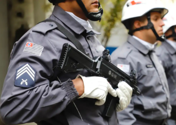 Policiais militares de São Paulo em uniforme cinza com capacete e armamento segurando arma de fogo preta