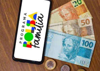 Smartphone exibindo o logotipo do Programa Bolsa Família na tela, posicionado sobre uma mesa de madeira ao lado de notas de 20, 50 e 100 Reais e moedas.