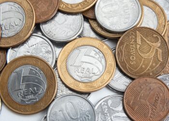 Diversas moedas do Plano Real espalhadas, incluindo moedas de 1 Real, 10 centavos e 25 centavos de diferentes anos; em evidência, uma moeda de 1 Real de 2008 com anel dourado e núcleo prateado.