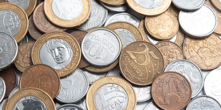 Close-up de uma mão segurando uma lupa sobre diversas moedas de um real e centavos espalhadas; a lupa destaca detalhes do cunho e possíveis erros de fabricação.