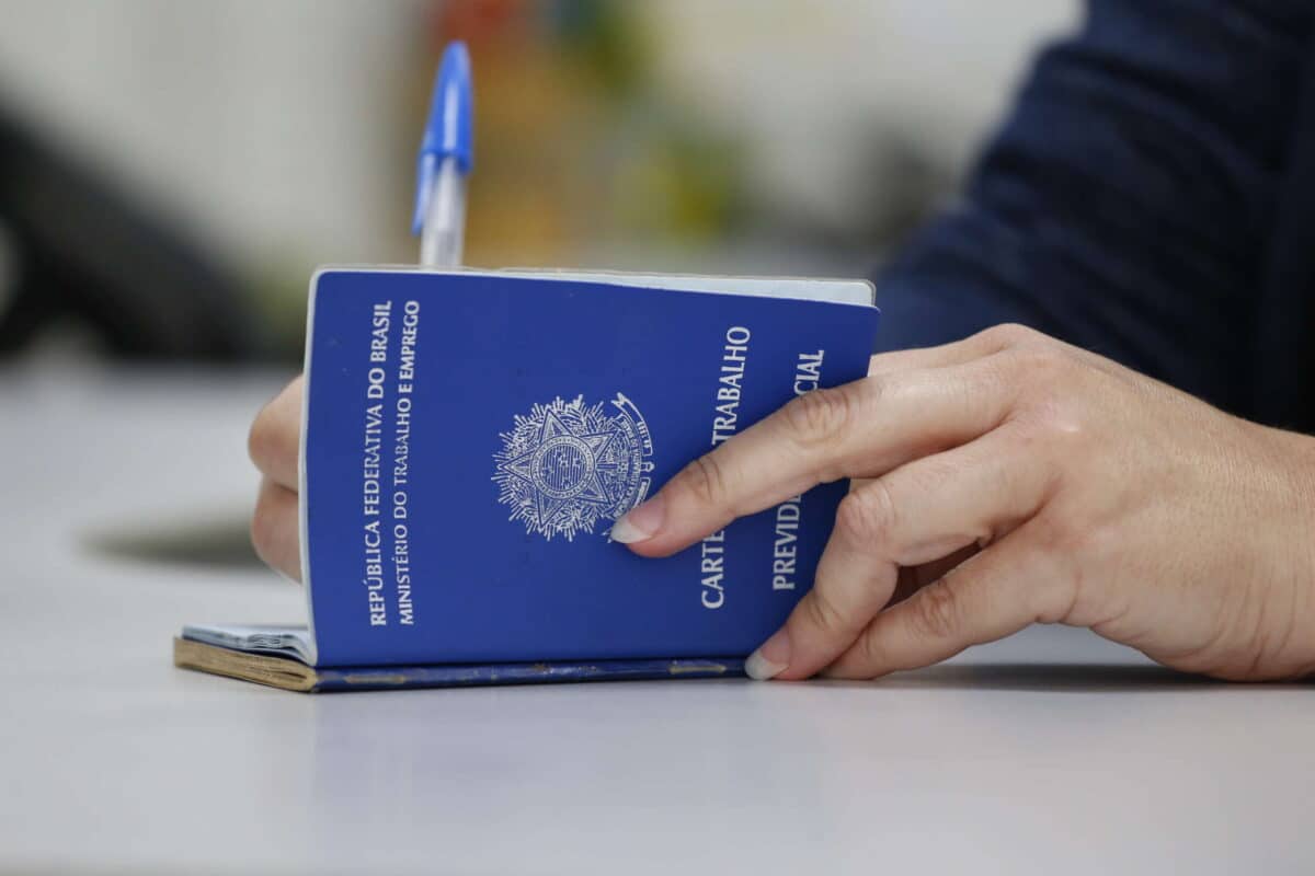 Emprego 2026 Mão feminina segurando carteira de trabalho brasileira azul com caneta azul ao fundo