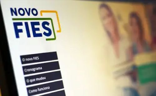 Tela de um computador mostrando o site oficial com o logotipo "NOVO FIES" em azul e verde. No canto inferior esquerdo, há botões de menu pretos com os textos: "O novo FIES", "Cronograma", "O que mudou" e "Como funciona". Ao fundo, de forma desfocada, aparecem estudantes sorrindo.