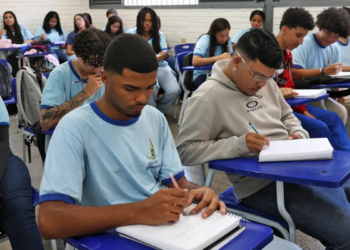 Jovens estudantes sentados em carteiras azuis escrevendo em cadernos em uma sala de aula com paredes de tijolos brancos