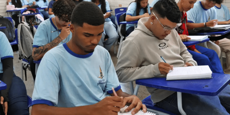 Jovens estudantes sentados em carteiras azuis escrevendo em cadernos em uma sala de aula com paredes de tijolos brancos