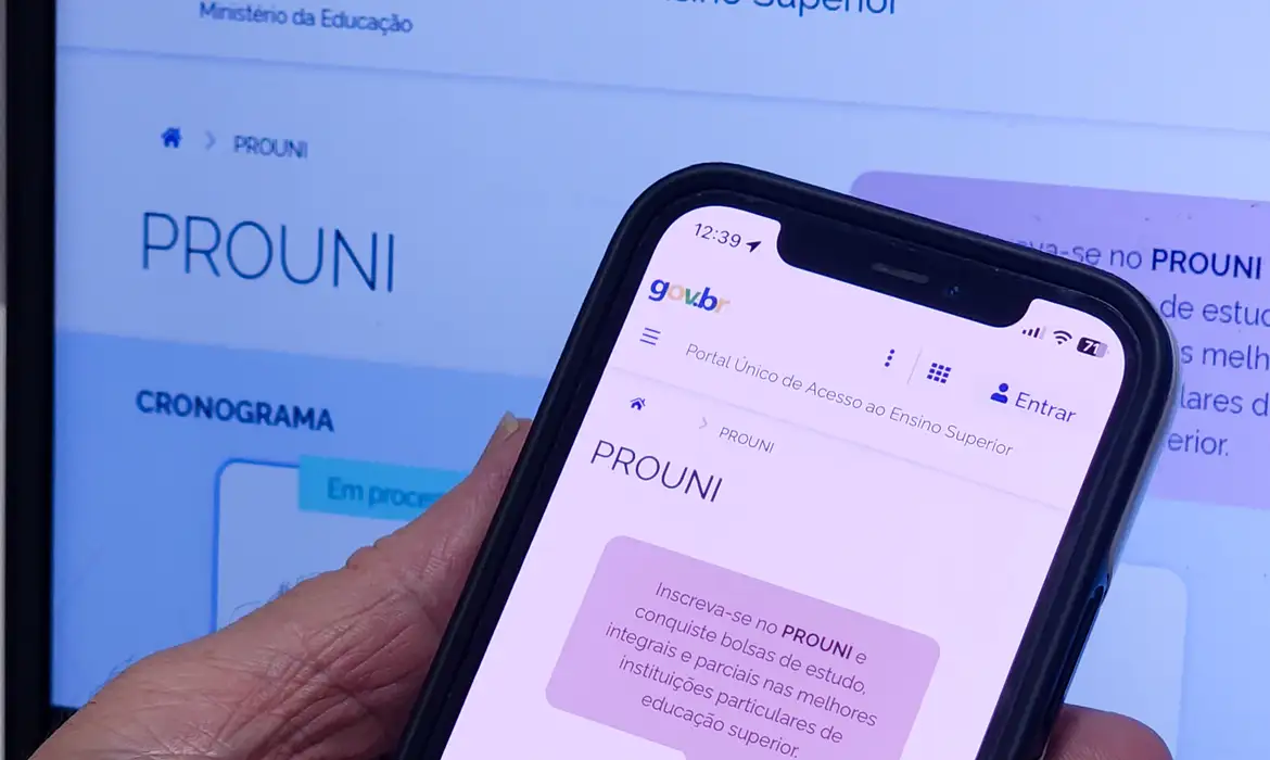 Inscrições para o prouni Mão segura smartphone exibindo o portal do Prouni, com a mesma tela no notebook ao fundo.