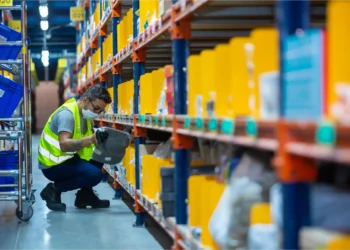 Funcionário em armazém do Mercado Livre, manuseando embalagem em prateleira de estoque, em um ambiente de logística e tecnologia.