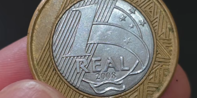 Close-up de uma moeda de 1 Real do ano de 2008 sendo segurada pelos dedos, destacando o valor e o ano gravados no centro prateado.