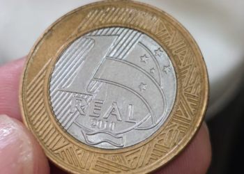 Close de uma mão segurando uma moeda de 1 real do ano 2010 entre os dedos. A moeda apresenta o disco central prateado com a palavra "REAL" e o ano, e o anel externo dourado com grafismos indígenas.