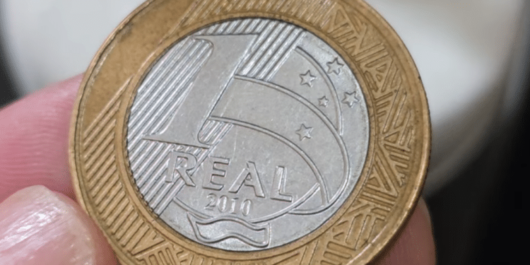 Close de uma mão segurando uma moeda de 1 real do ano 2010 entre os dedos. A moeda apresenta o disco central prateado com a palavra "REAL" e o ano, e o anel externo dourado com grafismos indígenas.