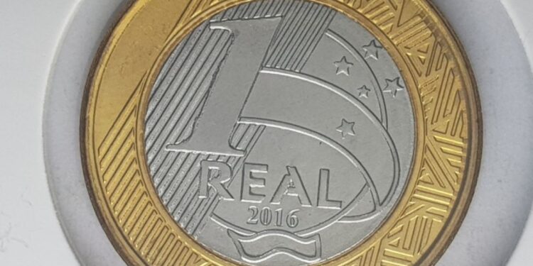 Close-up de uma moeda de 1 real de 2006 segurada por dedos. A imagem destaca a efígie da República no núcleo prateado e o anel dourado com a inscrição "BRASIL" e o ano "2006".