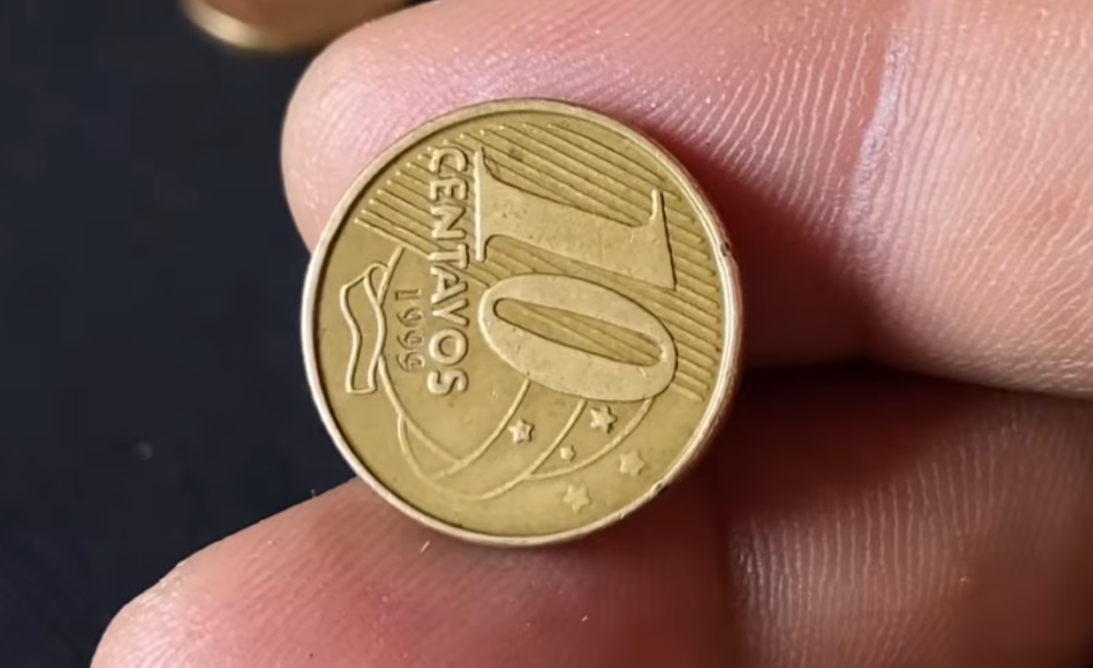 como identificar moeda reverso horizontal 1999 Close-up de uma mão segurando uma moeda de 10 centavos de Real do ano 1999. A moeda está rotacionada lateralmente para demonstrar o erro de cunhagem conhecido como reverso horizontal, onde o design aparece de lado em relação ao eixo vertical.