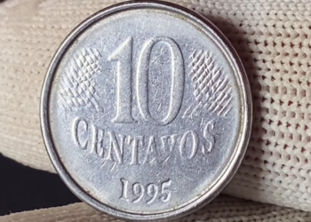 Close detalhado de diversas moedas brasileiras de um real e de centavos empilhadas sobre uma mesa de madeira, com foco em uma moeda de um real em destaque.