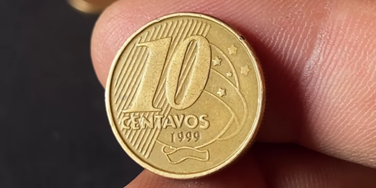Close-up de dedos segurando uma moeda de 10 centavos de Real, cor dourada, com o ano de 1999 visível na parte inferior. O fundo é escuro e desfocado.