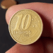 Close-up de dedos segurando uma moeda de 10 centavos de Real, cor dourada, com o ano de 1999 visível na parte inferior. O fundo é escuro e desfocado.