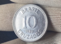 Close-up de uma moeda prateada de 10 Cruzados do ano de 1988, sendo segurada por uma mão com luva de tecido branco. A moeda exibe a inscrição "BRASIL", o valor "10" e "CRUZADOS" em destaque.