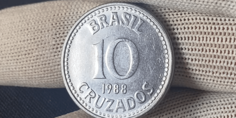 Close-up de uma moeda prateada de 10 Cruzados do ano de 1988, sendo segurada por uma mão com luva de tecido branco. A moeda exibe a inscrição "BRASIL", o valor "10" e "CRUZADOS" em destaque.