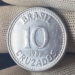 Close-up de uma moeda prateada de 10 Cruzados do ano de 1988, sendo segurada por uma mão com luva de tecido branco. A moeda exibe a inscrição "BRASIL", o valor "10" e "CRUZADOS" em destaque.