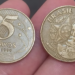 Duas moedas de 25 centavos de 1999 sendo seguradas, mostrando as faces anverso (busto de Deodoro) e reverso (valor "25 centavos").
