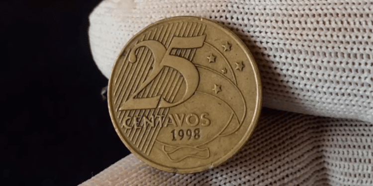 Close-up de uma moeda de 25 centavos de 1998 sendo segurada por dedos com luvas brancas, focando no reverso onde a data aparece marcada sob os ramos.