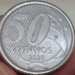 Fotografia em close-up do reverso de uma moeda de 50 centavos de 2020, segurada entre os dedos. A imagem destaca o número 50, a palavra "CENTAVOS" e o ano de fabricação 2020 sobre o fundo de linhas diagonais.
