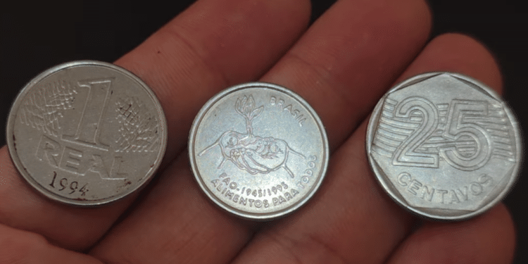 Três moedas brasileiras raras estendidas sobre a palma de uma mão: uma de 1 Real de 1994, uma comemorativa da FAO com o desenho de mãos segurando uma planta e uma de 25 centavos de 1995.