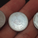 Três moedas brasileiras raras estendidas sobre a palma de uma mão: uma de 1 Real de 1994, uma comemorativa da FAO com o desenho de mãos segurando uma planta e uma de 25 centavos de 1995.