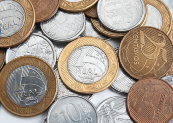 Uma pilha mista de moedas do Real brasileiro de diversos valores, incluindo moedas de 1 Real, 25 centavos, 10 centavos e 5 centavos, espalhadas sobre uma superfície clara.