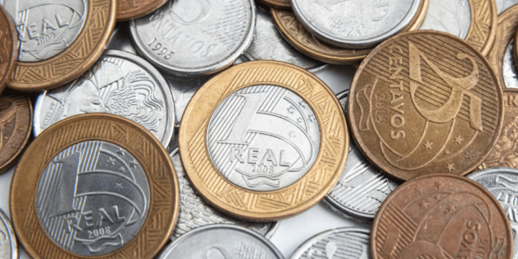 Uma pilha mista de moedas do Real brasileiro de diversos valores, incluindo moedas de 1 Real, 25 centavos, 10 centavos e 5 centavos, espalhadas sobre uma superfície clara.