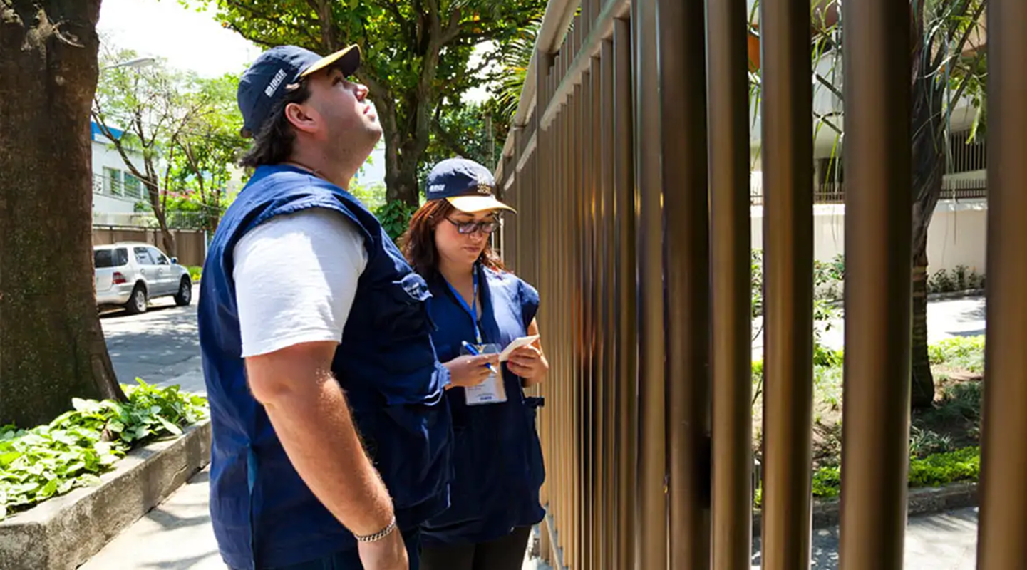 Principais datas do cronograma do concurso do IBGE Recenseadores do IBGE com colete e boné azul realizando coleta de dados em área residencial.