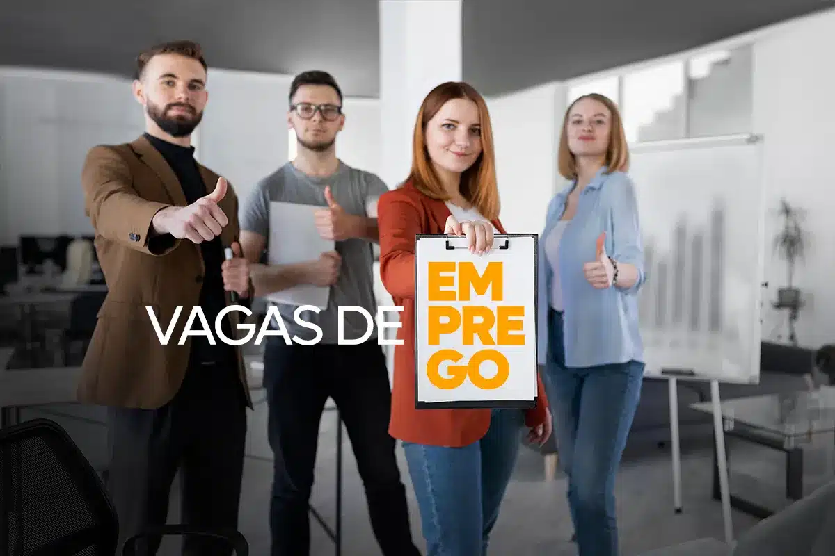 Grupo de quatro profissionais jovens em um escritório moderno fazendo sinal de "joinha". A mulher ao centro segura uma prancheta branca com as palavras "VAGAS DE EMPREGO" escritas em preto e laranja.