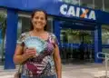 Mulher segurando cédulas de dinheiro brasileiro em frente a uma agência da Caixa representando os pagamentos do Bolsa Família de julho.