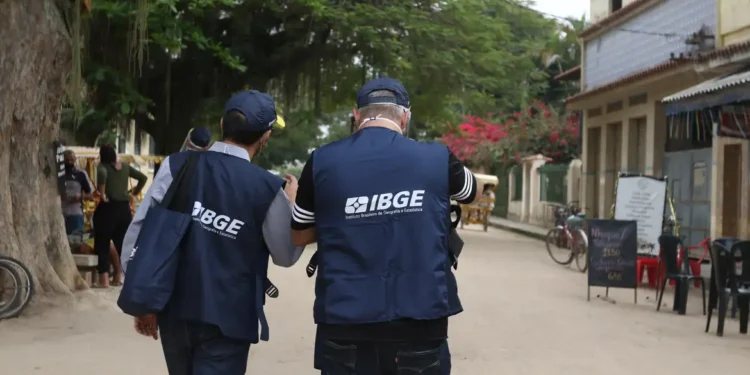 Dois recenseadores do IBGE com coletes azuis caminhando em rua com árvores, lojas e bicicletas